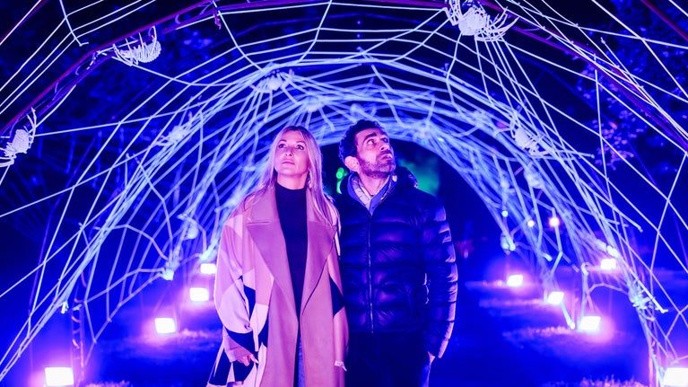 A man and a woman with frightened expressions walking through a tunnel adorned with large white spiders and their webs, illuminated by unsettling purple lights